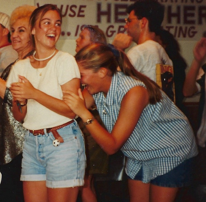 Heather Martin, who later passed away from cervical cancer in her twenties, shares a joyful moment with her best friend Sara Lyle-Ingersoll, now Communications Director for Cervivor, Inc. In this candid photo from their senior year graduation event, Heather, wearing a white t-shirt and denim shorts, laughs with Sara, who is playfully leaning on her shoulder, dressed in a sleeveless striped shirt and shorts. They are surrounded by other attendees in a festive atmosphere.