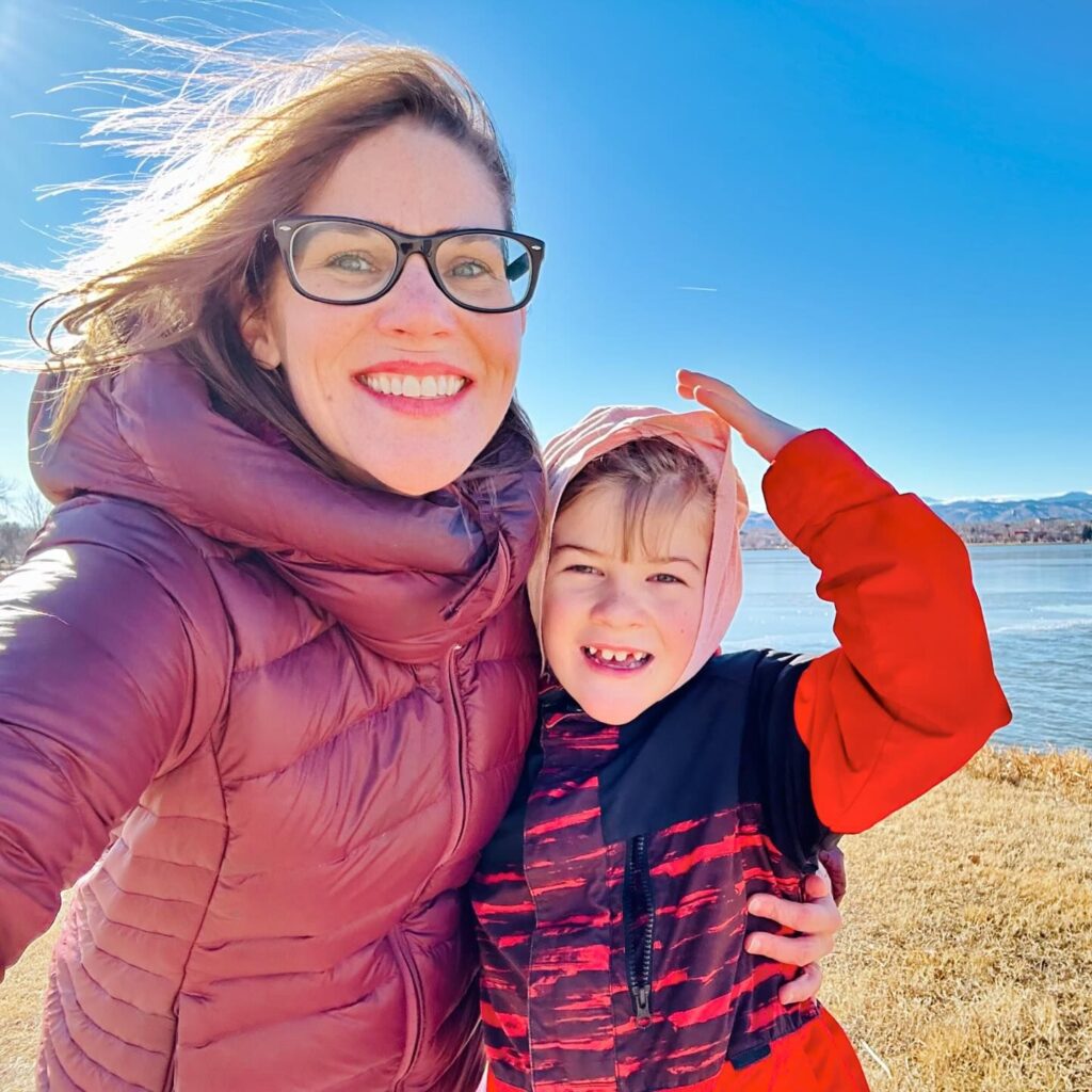 Sara Lyle-Ingersoll smiles brightly as she poses with her 9-year-old son, Loch, outdoors in Denver during the winter. They are bundled up in winter jackets, with Sara wearing a purple puffer coat and Loch in a red and black jacket. The sky is clear and blue, and the mountains are visible in the background.