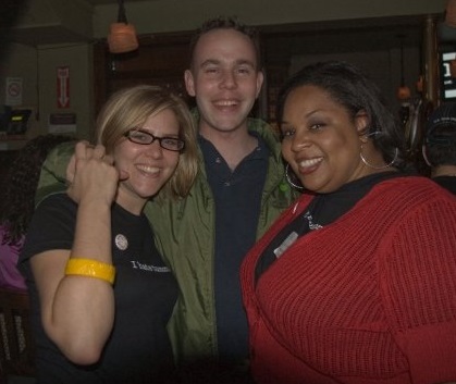 Sara Lyle-Ingersoll, wearing an "I Hate Tumors" shirt, stands with a man and Tamika Felder at a cervical cancer awareness event in 2009. Sara is smiling and holding up her wrist, showcasing a yellow bracelet, while Tamika, dressed in a red cardigan, stands on the right, also smiling.