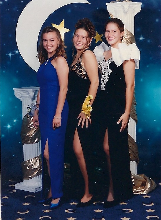 Heather, Mindy, and Sara Lyle-Ingersoll stand together at their senior prom in the 1990s. Sara, on the right, is wearing a black gown with a large white bow on the shoulder. Mindy is in the center wearing a black dress with gold accents, and Heather is on the left in a blue dress. They pose in front of a backdrop featuring a crescent moon, stars, and decorative columns.