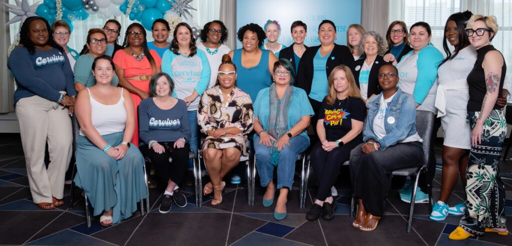 A diverse group of individuals, all attendees of the 2024 Cervivor School, pose for a group photo in a well-lit room decorated with teal and white balloons. The group consists of women wearing Cervivor-themed shirts, casual and professional attire, with a warm, supportive atmosphere. Some attendees are seated while others stand behind them.