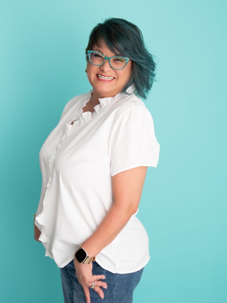 This image shows cervical cancer survivor, Christy, who is from North Carolina. She has teal hair and glasses with a ruffled white shirt.