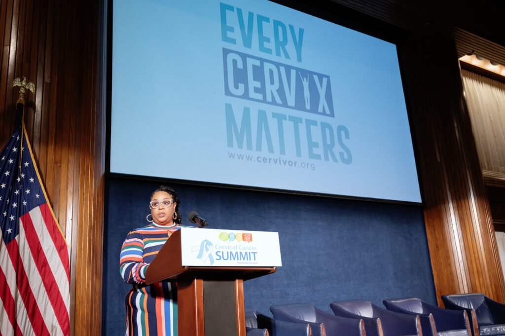 Tamika Felder, Founder and Chief Visionary of Cervivor, welcomes attendees at the 2025 Cervical Cancer Summit during her annual State of Cervical Cancer speech. She wears a long-sleeve, colorful striped dress, glasses, and a long braided ponytail while speaking at the lectern.