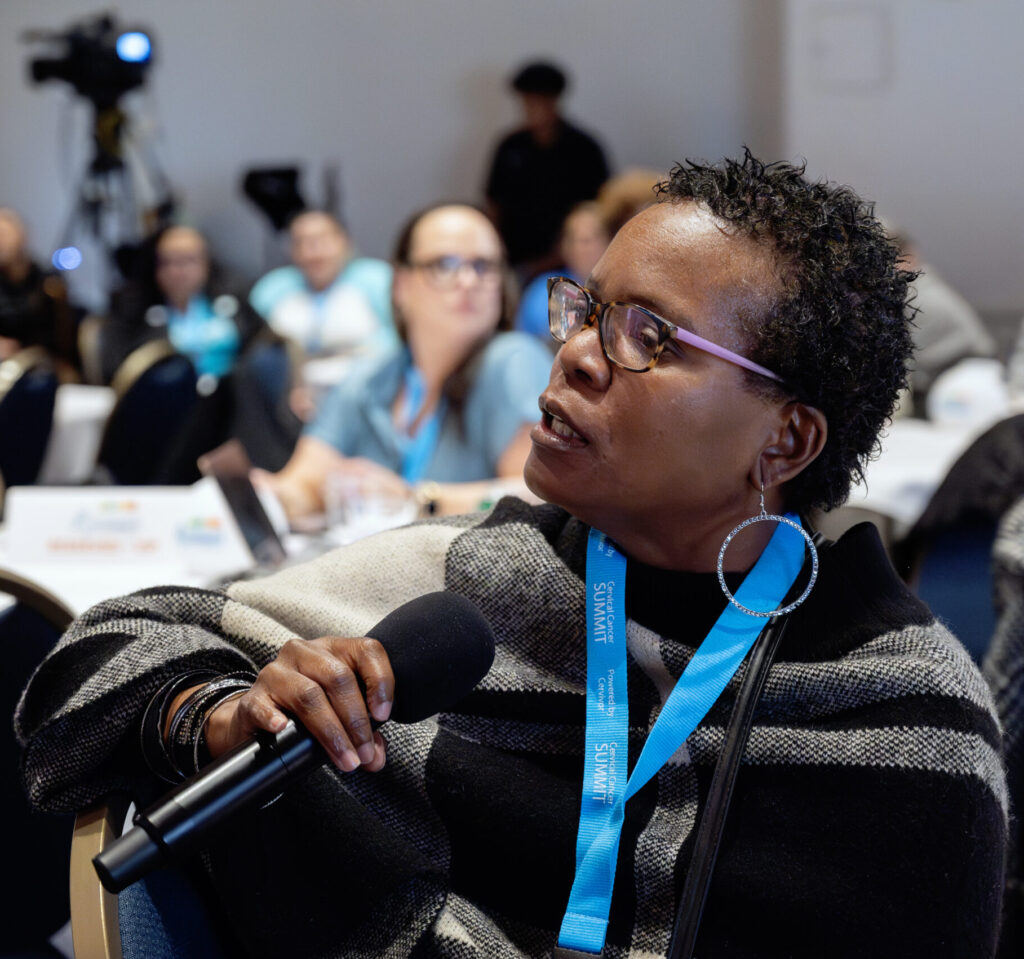 Gwen Jackson, wearing a black and white plaid sweater and a Summit lanyard, holds a microphone while asking a question from the audience at the 2025 Cervical Cancer Summit.