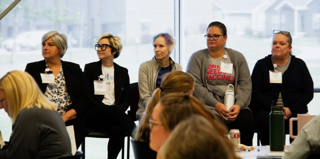 The image depicts a group of women seated in a conference room, with five women prominently featured on the right side of the frame. They are all wearing name tags and appear to be engaged in a discussion or presentation.
The women are dressed in casual attire, with some wearing blazers or cardigans over their shirts.
The woman on the far left has short gray hair and is wearing a black blazer over a white blouse with a floral pattern.
The second woman from the left has short blonde hair and is wearing a black blazer over a white t-shirt with blue and red writing.
The third woman from the left has short blonde hair with multicolored highlights and is wearing a gray cardigan over a dark blue shirt.
The fourth woman from the left has dark hair pulled back and is wearing a gray cardigan over a gray t-shirt with red writing that reads "IOWA NEEDS VACCINES."
The woman on the far right has dark hair pulled back and is wearing a black hoodie over a gray shirt.
In the foreground, several other women are seated at tables, facing the five women on the right.
The background of the image features large windows that allow natural light to enter the room and provide a view of the outside environment.
The overall atmosphere of the image suggests a professional or educational setting, possibly a conference or workshop.