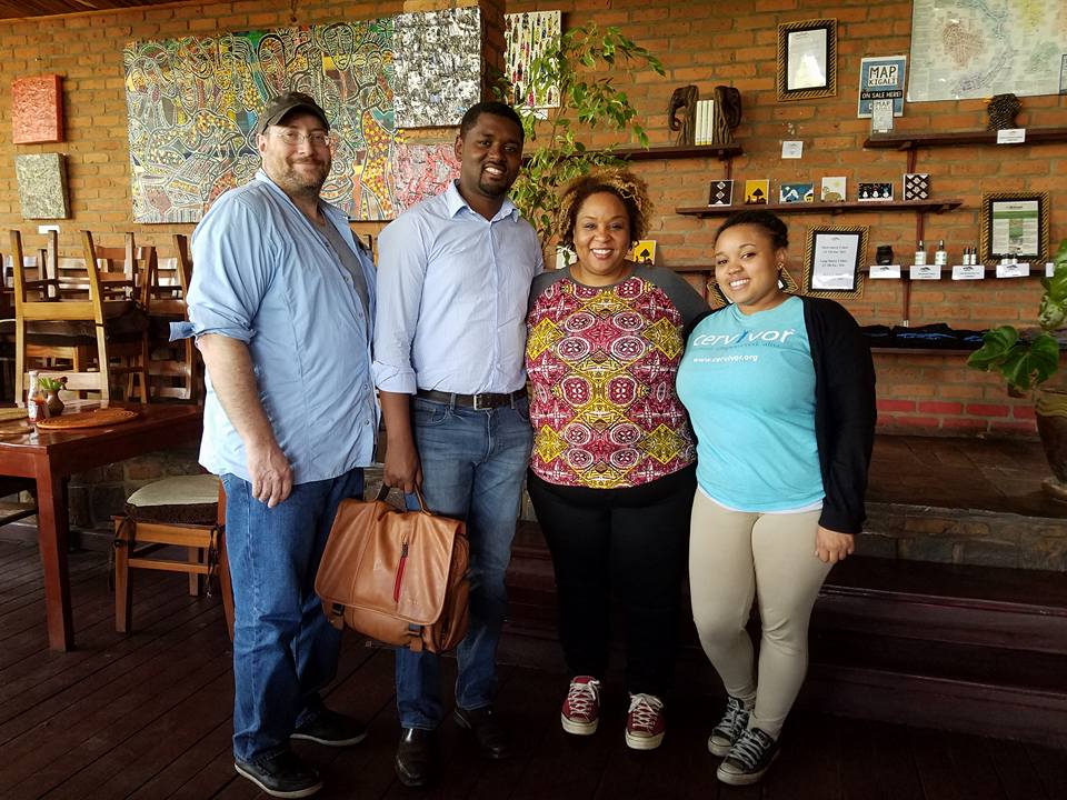 The image shows four people standing together in a restaurant or cafe. The man on the left is wearing a light blue shirt and jeans, with a black cap. The man next to him is wearing a light blue button-down shirt and jeans, holding a brown leather bag. The woman beside him has curly hair and is wearing a colorful patterned top with black pants and red sneakers. The woman on the right is wearing a light blue t-shirt with a black cardigan and khaki pants. The background features a brick wall adorned with various paintings and decorations, including a large abstract piece behind the group. The atmosphere appears to be casual and friendly, suggesting that the individuals are friends or acquaintances gathered for a social occasion.