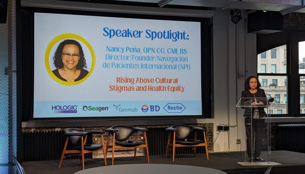 The image depicts a woman standing at a podium on a stage, addressing an audience. The stage is set with a large screen behind her, displaying a presentation slide titled "Speaker Spotlight" in blue text. Below the title, a circular photo of the speaker, Nancy Peña, is accompanied by her name and credentials in blue text, as well as her title and organization in smaller blue text. The slide's subtitle, "Rising Above Cultural Stigmas and Health Equity," is written in red text.