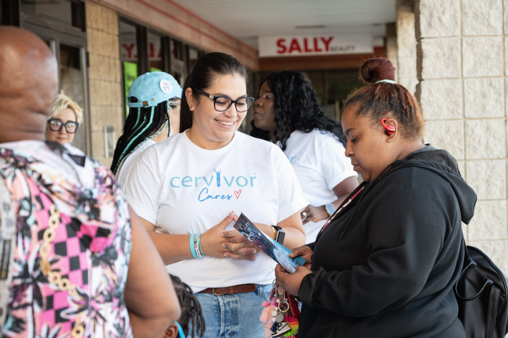 The image depicts a group of people gathered outside a storefront, with one woman prominently wearing a white t-shirt featuring the text "cervivor Cares" in blue and red. The scene appears to be a promotional or awareness event, possibly related to cervical cancer or women's health.
Key Elements:
A woman in the center of the image is wearing a white t-shirt with the text "cervivor Cares" printed on it.
She is holding a pamphlet and engaging in conversation with another woman.
The surrounding individuals are also dressed casually, suggesting an informal gathering.
The storefront behind them features a sign reading "SALLY Beauty," indicating that the event is taking place outside a beauty supply store.