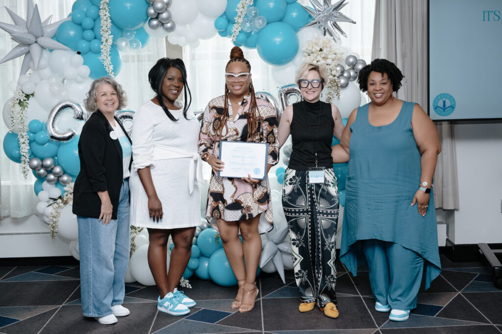 The image depicts a group of five women standing together, posing for a photograph in front of a vibrant balloon backdrop. The women are dressed in various outfits, with the central figure holding a certificate.
The woman on the far left is attired in a black cardigan and blue jeans.
The second woman from the left wears a white dress.
The third woman, positioned centrally, holds a certificate and is dressed in a patterned dress.
The fourth woman from the left is wearing a black top and patterned pants.
The woman on the far right is dressed in a teal dress.
The background features a wall adorned with balloons in shades of white, blue, and silver, accompanied by white flowers. A projector screen is visible on the right side of the image, displaying a logo and the text "IT'S GRADUATION TIME!" in blue letters. The floor is composed of dark tiles arranged in a geometric pattern.
The overall atmosphere of the image suggests that the women are celebrating a special occasion or achievement, as indicated by the certificate being held by the central figure.