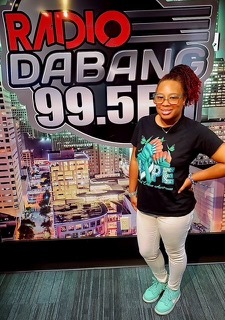 The image shows a woman standing in front of a large sign that reads "RADIO DABANG 99.5 FM."
Here's what's in the image:
The woman is wearing:
A black graphic t-shirt with a colorful design and the word "HOPE" in teal letters
White pants
Teal sneakers
Her hair is styled in a short, curly updo with red highlights
She's smiling and has her left hand on her hip
The sign behind her features:
A cityscape at night with many lit-up buildings
The words "RADIO DABANG 99.5 FM" in large letters, with "RADIO" in red and the rest in white and gray
The floor is made of gray carpeting with a striped pattern
The image appears to be a promotional photo for a radio station, possibly taken in a studio or office. The woman may be a DJ, host, or representative of the station.