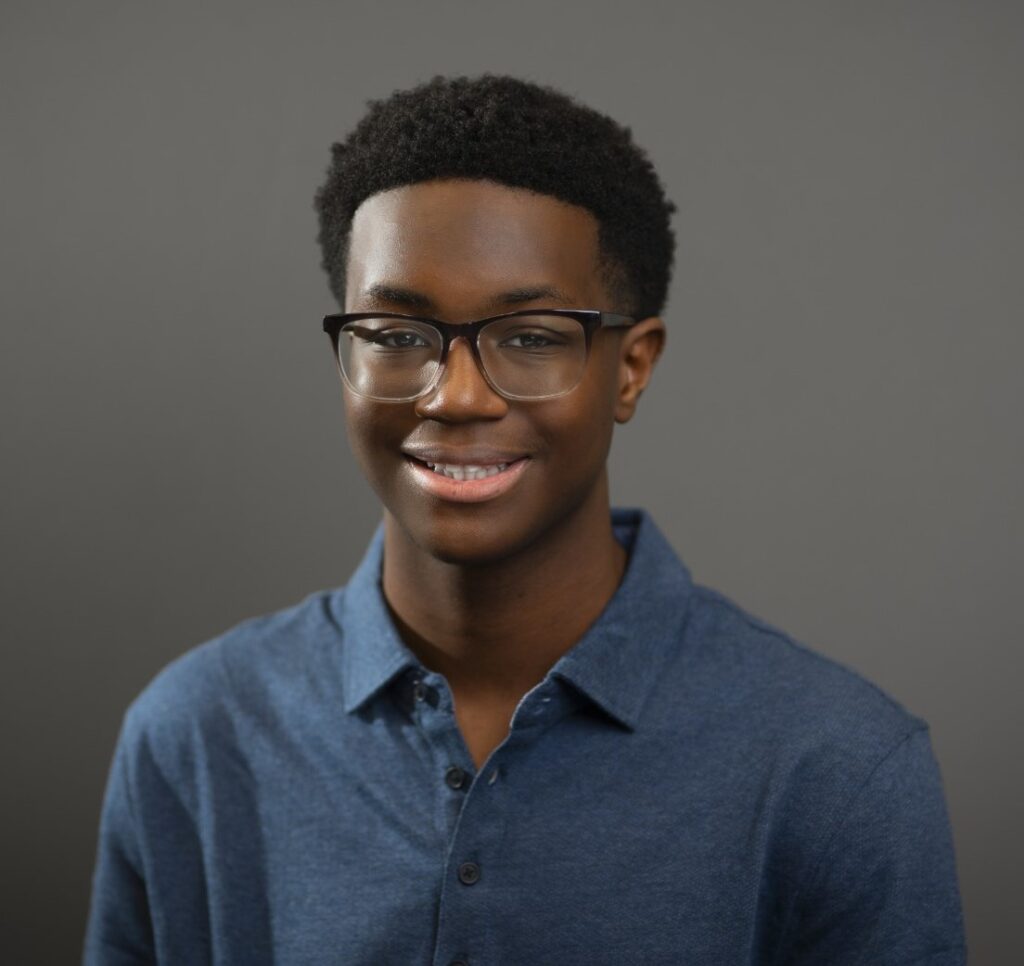The image is a portrait of a young man with dark skin and short, curly black hair. He is wearing a blue collared shirt and black-framed glasses, and is smiling at the camera.
The man has dark skin and short, curly black hair.
His hair is cut close to his head, with a slight fade at the sides.
His eyebrows are thick and well-groomed.
He is wearing a blue collared shirt.
The shirt is a medium blue color and appears to be made of a lightweight material.
It has a relaxed fit and is buttoned up to the top.
He is also wearing black-framed glasses.
The frames are rectangular in shape and have a subtle curve at the temples.
The lenses are clear and do not appear to have any tint or coating.
The man is smiling at the camera.
His smile is wide and genuine, showing off his white teeth.
His eyes are crinkled at the corners, giving him a friendly and approachable appearance.
The background of the image is a plain gray color.
The gray is a medium tone, neither too light nor too dark.
It provides a neutral backdrop that allows the subject to stand out.
Overall, the image presents a friendly and approachable young man who appears to be confident and comfortable in front of the camera