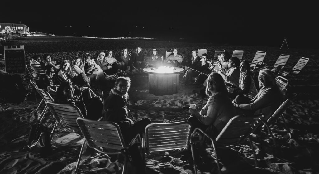 Group of Cervivor School attendees sitting in a circle around a fire pit on a beach at night, sharing support and connection